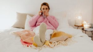 Woman in a pink sweater relaxing in bed with a warm drink, surrounded by soft blankets and candles.