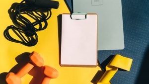 Flat lay of fitness equipment including dumbbells, jump rope, and clipboard on a yellow background.